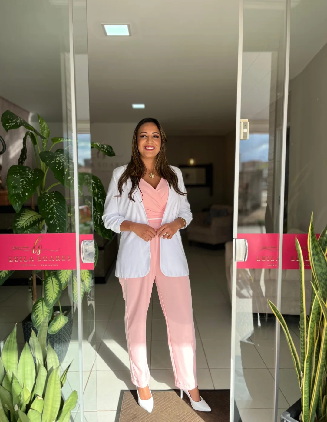 Leila Soares sorrindo na porta de vidro do centro estético, de blazer branco e conjunto rosa, entre vasos de plantas