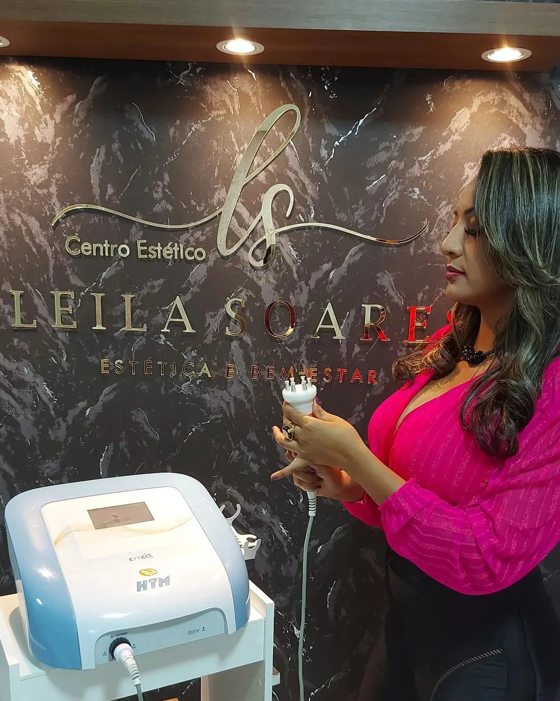 Leila segurando a manopla do aparelho de radiofrequência, diante de uma parede escura com o nome do centro estético em dourado