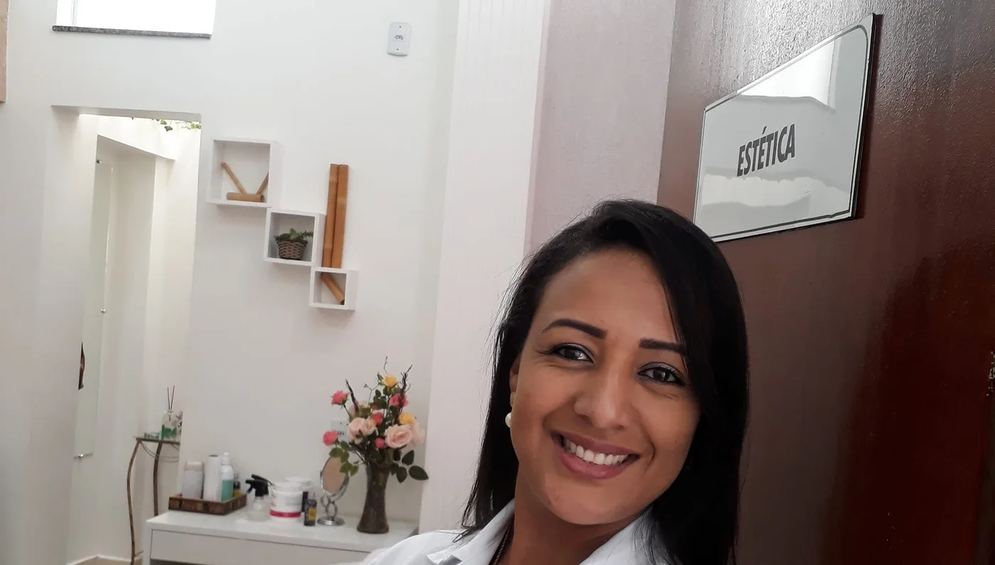 Leila de jaleco branco, sorrindo, ao lado de uma porta com a placa ESTÉTICA